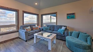 A living room with a gray sofa, a white coffee table, and a view of the beach through large windows. - Sanderling Sea Cottages, Unit 7 w/ Beach Access! (Waldport)