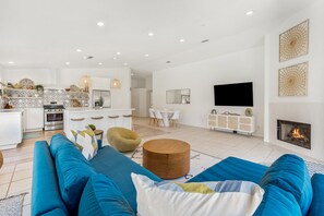 Una sala de estar moderna con un sofá azul, un taburete redondo de madera y un comedor con sillas blancas y una mesa.