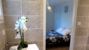 Un baño con un lavamanos blanco y un jarrón con orquídeas blancas. Hay una zona de ducha con ducha y toallero. El baño tiene azulejos color beige y una puerta blanca.