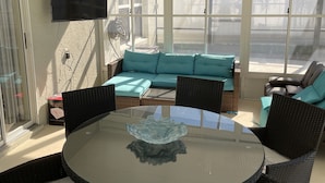 Eine sonnendurchflutete Terrasse mit einem runden Glastisch, Korbstühlen, einem blaugrünen Sofa und einem an der Wand befestigten Fernseher.