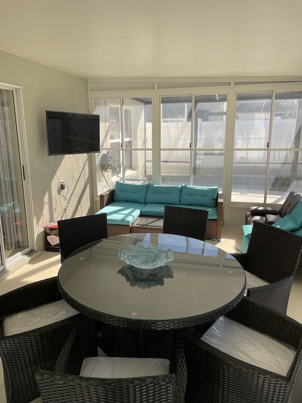 Une terrasse ensoleillée avec une table ronde en verre, des chaises en osier, un canapé de couleur turquoise et un téléviseur fixé au mur.
