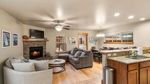 A spacious living room with a fireplace, a grey sofa, a coffee table, and a kitchen area with a granite countertop and wooden cabinets. - Charming Home by the Park with a Hot Tub (Bozeman)