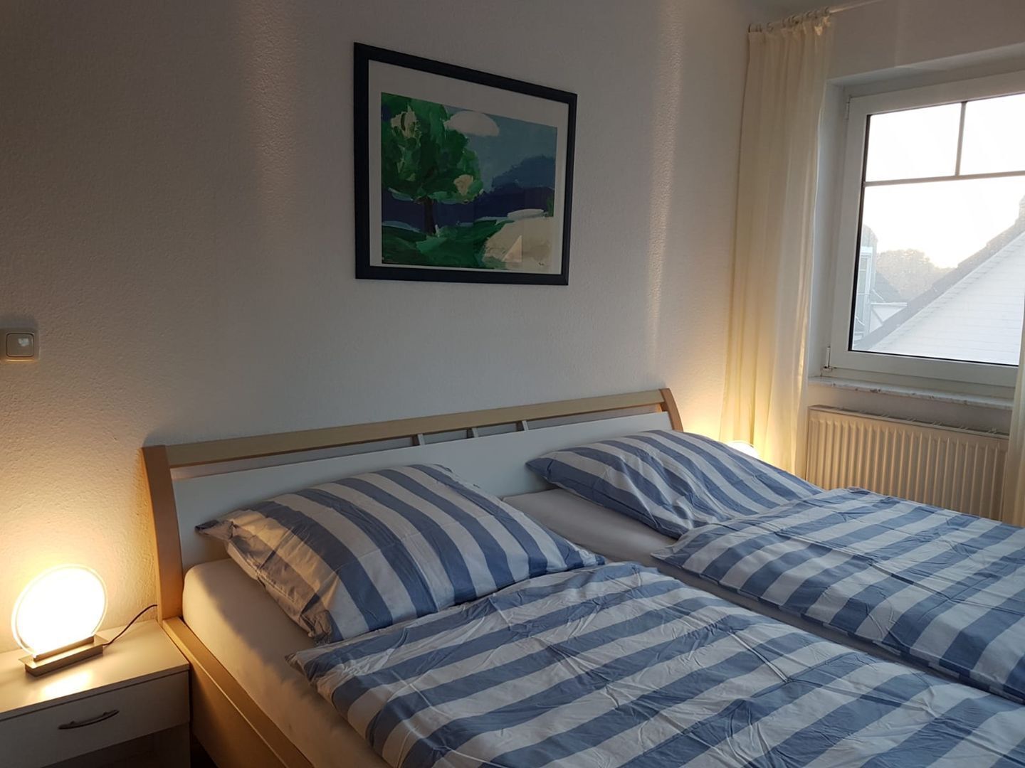 A bedroom with a bed, a bedside table with a lamp, a framed picture on the wall, and a window with curtains.