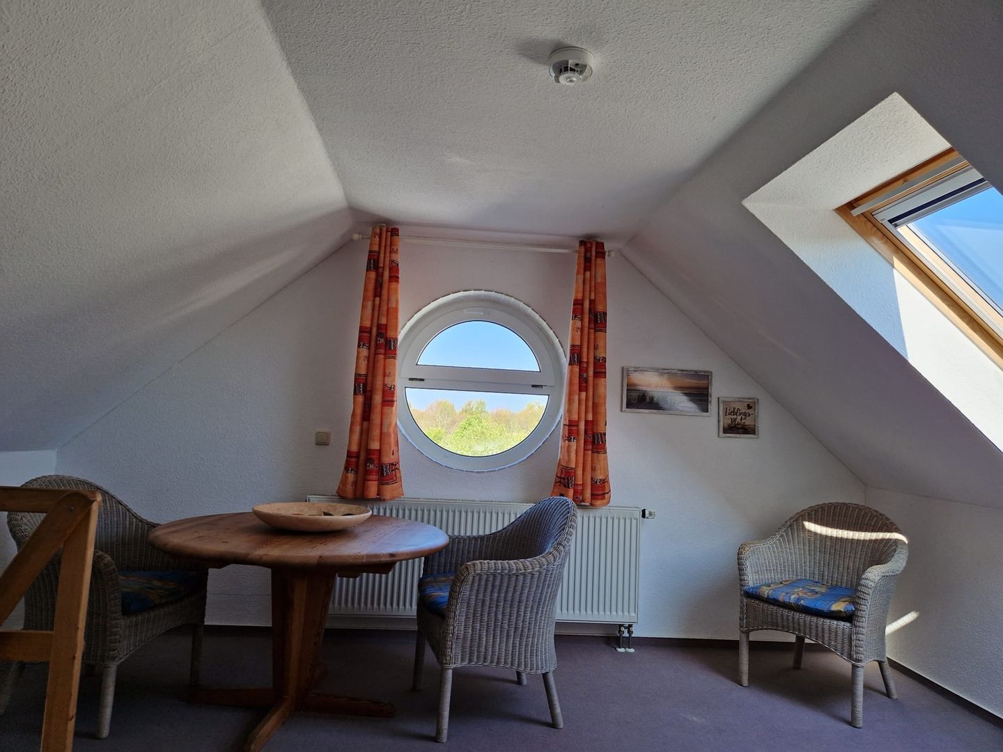 A cozy attic room with a round window, two wicker chairs, a wooden table, and a radiator.