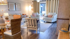 A spacious living room with a large window, a ceiling fan, and a dining area with wicker furniture. - Tipsy Turtle Edgewater Villa 2901-Pickleball, Tennis, Fitness Center Included! 1st Floor-End Unit (Panama City Beach)