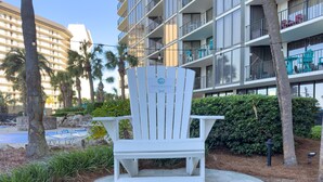 A white Adirondack chair in a landscaped area with a pool and palm trees. - Tipsy Turtle Edgewater Villa 2901-Pickleball, Tennis, Fitness Center Included! 1st Floor-End Unit (Panama City Beach)