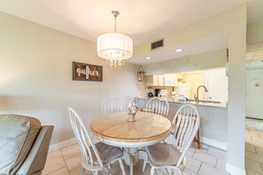 A dining area with a round table, white chairs, and a chandelier. There is a kitchen in the background with white cabinets and a sink.