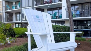 Una silla lounge blanca con un letrero que dice 'EDGEWATER' en un área ajardinada con un camino empedrado, rodeada de follaje y un edificio de varios pisos con balcones.