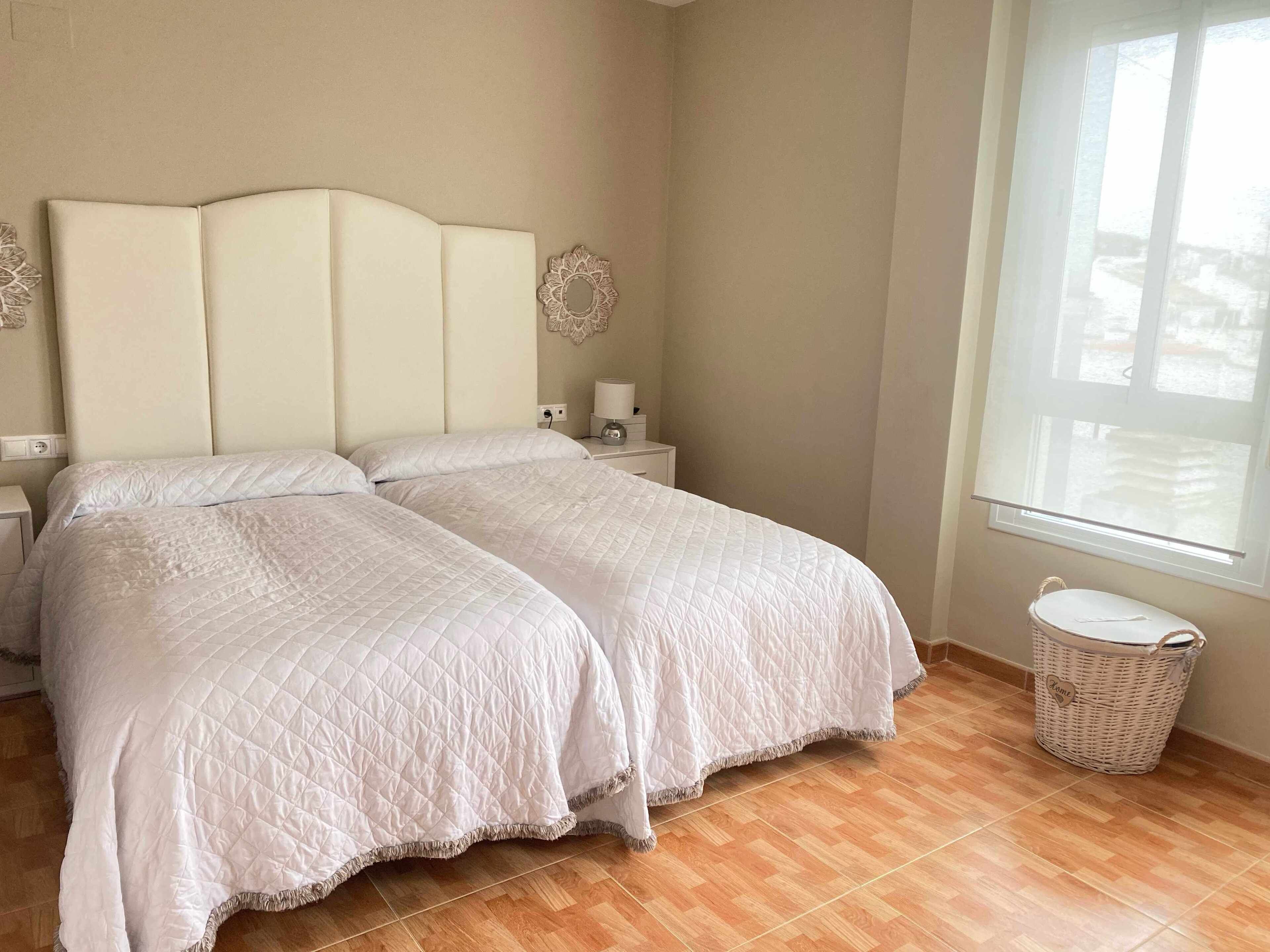 Una cama doble con edredón blanco acolchado, una mesita de noche con lámpara, un adorno en la pared y una cesta de mimbre junto a la ventana.