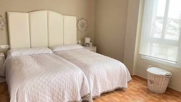 Una cama doble con edredón blanco acolchado, una mesita de noche con lámpara, un adorno en la pared y una cesta de mimbre junto a la ventana.