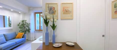 A modern living room with a blue sofa, a wooden coffee table, and two vases with white flowers. There are two framed artworks on the wall and a door in the background.