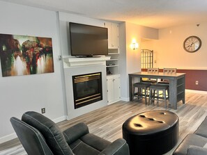 Un salon moderne doté d’une cheminée, d’un téléviseur à écran plat, d’un coin repas avec une table et des chaises, et d’une horloge murale.