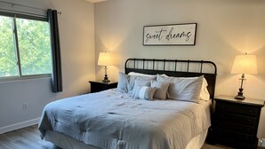 Une chambre à coucher avec un grand lit, deux tables de chevet avec des lampes, une fenêtre avec des rideaux et un tableau accroché au mur.