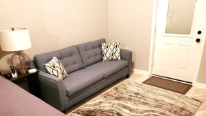 Una habitación con un sofá gris, un tapete estampado, una mesita de noche con una lámpara y una puerta con felpudo.