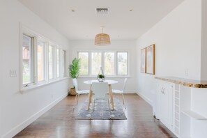 Uma cozinha clara com armários brancos, uma mesa de jantar redonda com cadeiras, uma luminária pendente e janelas amplas.