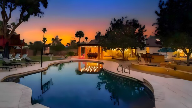 Una piscina con sillas de descanso y un área de estar cubierta, rodeada de árboles y un atardecer al fondo.