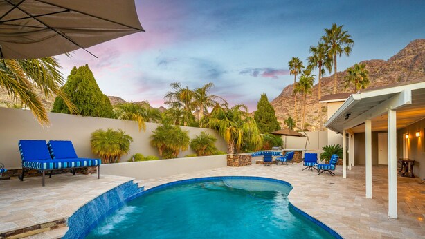 A swimming pool with blue-tiled edges, surrounded by lounge chairs and palm trees, set against a mountainous backdrop. - Biltmore Highlands by Porter | Luxury Home w/Private Pool and Mountain Views (Phoenix)