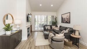 A modern living room with a sectional sofa, a fireplace, a television, and a large window with blinds.