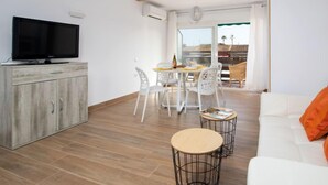 Uma sala de estar moderna com uma televisão de tela plana, um armário de madeira, uma área de refeições com cadeiras brancas e um sofá branco com almofada laranja.
