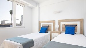 Quarto de hotel com duas camas, cada uma com cabeceira de madeira e almofada azul. Há uma janela com vista para palmeiras lá fora.