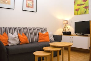 Un salon avec un canapé à rayures, des coussins orange, une table basse en bois, une lampe et un téléviseur posé sur un meuble.