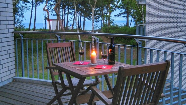 A wooden table with a candle, wine glasses, and a bottle on a balcony with a view of trees and a building in the background.