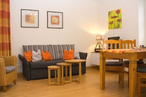 A living room with a sofa, two side tables, a dining table with chairs, a lamp, and two framed artworks on the wall.