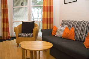 A living room with a wicker chair, a dark grey sofa with orange pillows, and a round wooden table.