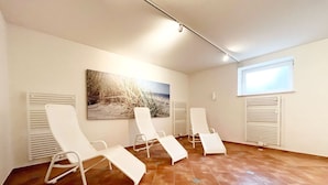 A room with four white lounge chairs, a large painting of a beach scene, and a window with blinds. - Dünenresidenz - Apartment directly on the beach, balcony with sea view - Dünenresidenz FeWo (Breege)
