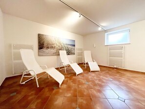 A room with four white lounge chairs, a large painting of a beach scene, and a window with blinds. - Dünenresidenz - Apartment directly on the beach, balcony with sea view - Dünenresidenz FeWo (Breege)