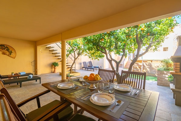 A dining area with a table set for four, surrounded by chairs, with a view of a tree and a building in the background.