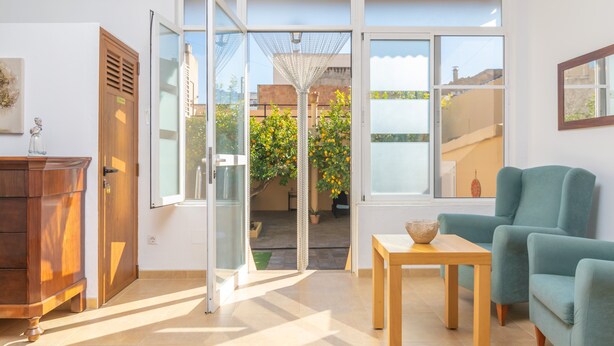A modern living room with a wooden cabinet, a teal armchair, a small wooden table, and a glass door leading to an outdoor area with a potted plant and a view of buildings. - JUNIPER (INCA) - Cozy town house with above ground pool in the center of Mallorca. Free WI (Inca)