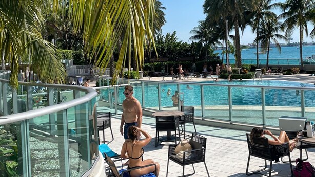 Un espace aménagé au bord de la piscine, avec des chaises longues, une rambarde en verre et des palmiers.