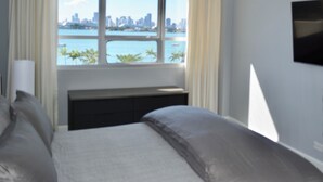 Une chambre avec une grande fenêtre donnant sur le panorama urbain et l’étendue d’eau.