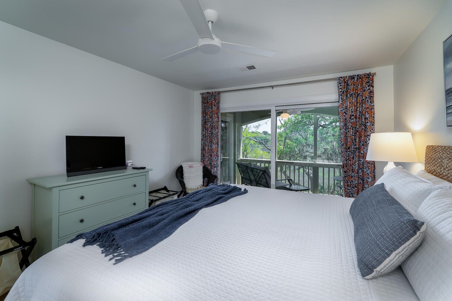 Una habitación de hotel con cama, televisor en una cómoda, balcón con cortinas y ventilador de techo.