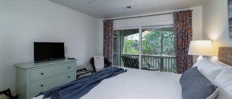 Una habitación de hotel con cama, televisor en una cómoda, balcón con cortinas y ventilador de techo.