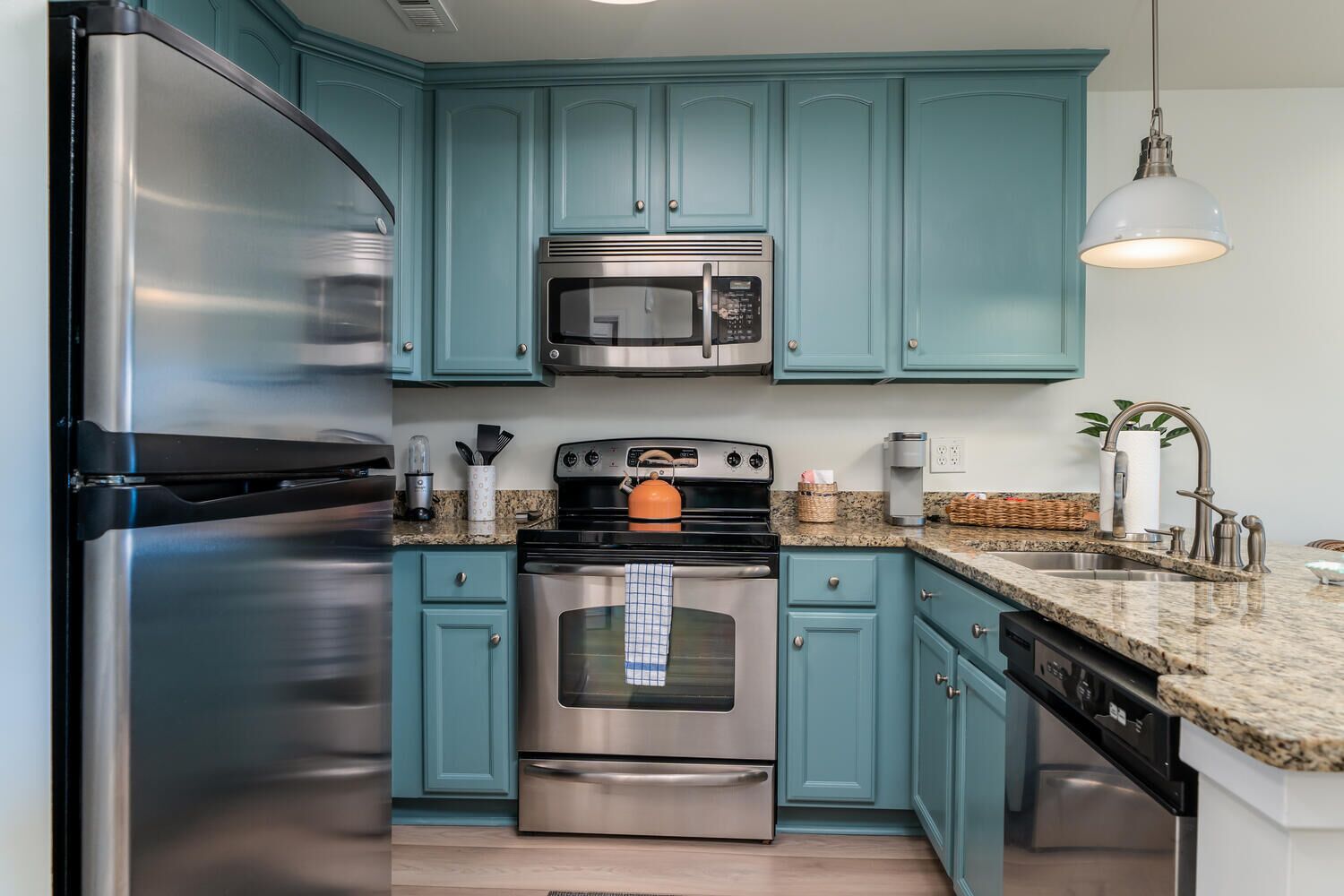 A kitchen with teal cabinets, stainless steel appliances, granite countertops, and a hanging light fixture.