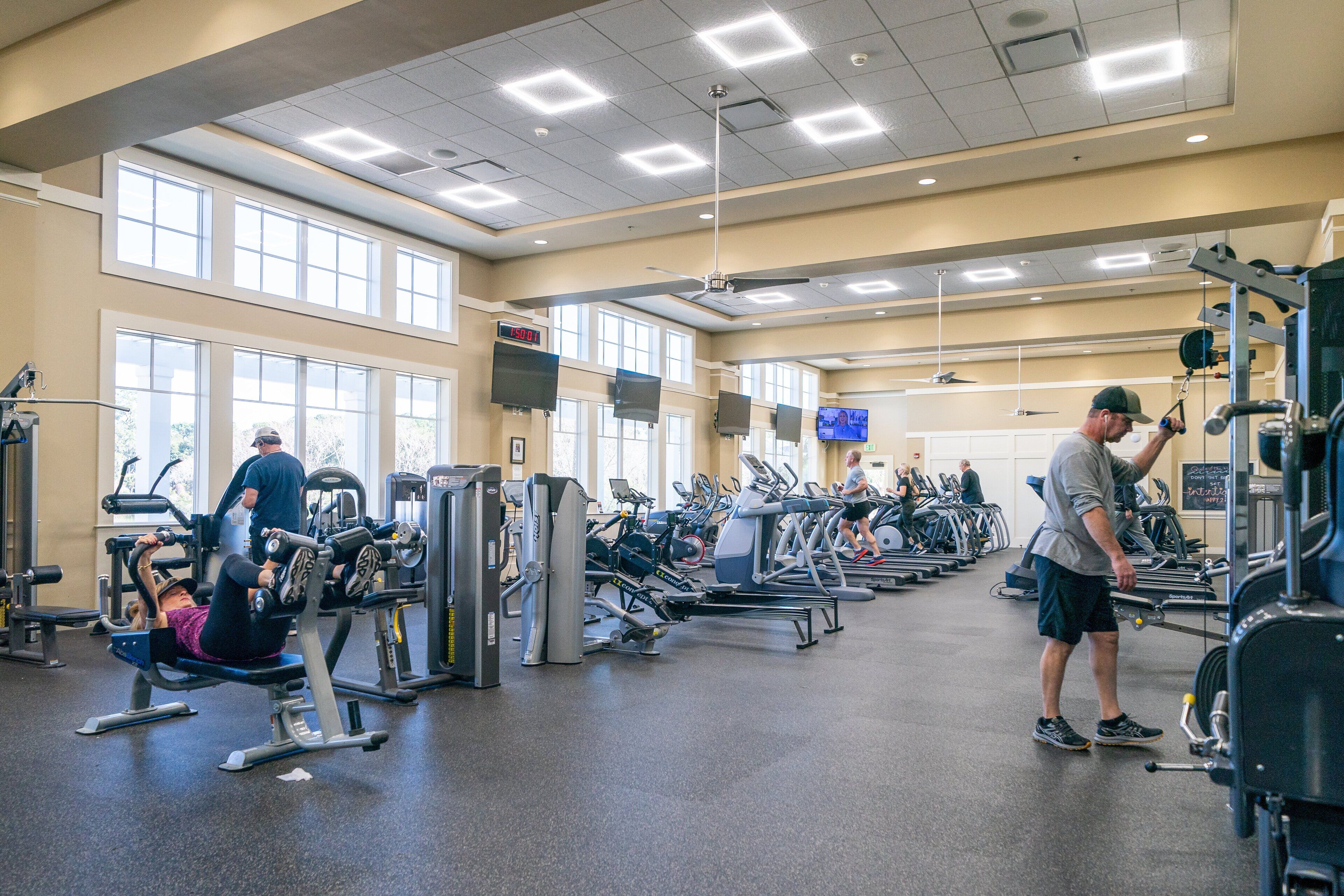 Ein Fitnessstudio mit unterschiedlichen Trainingsgeräten, darunter Laufbänder und Kraftgeräte, und großen Fenstern für natürliches Licht.