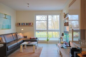 A modern living room with a leather sofa, a coffee table, and a TV. There is a bookshelf with books and a large window with a view of the outdoors.