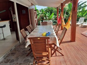 Un espace de restauration en plein air avec une longue table et des chaises en bois, entouré d’une terrasse en bois et d’une végétation luxuriante.