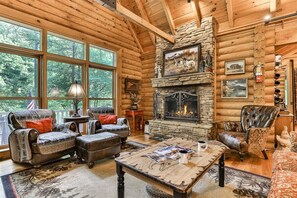 L’intérieur d’une cabane en rondins, avec une cheminée en pierre, de grandes fenêtres et des meubles rustiques.