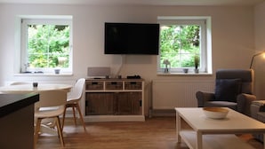 Un soggiorno moderno con una TV a schermo piatto montata a parete, pavimento in legno, mobili bianchi e ampie finestre con vista su del verde.