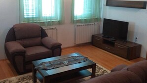A living room with a brown sofa, a brown armchair, a coffee table, a TV on a wooden stand, and two windows with green curtains. - Remarkable 3-Bed House in Zabljak (Zabljak)