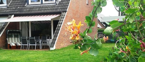 A residential backyard with a covered patio, a brick house, and a garden with flowering plants.