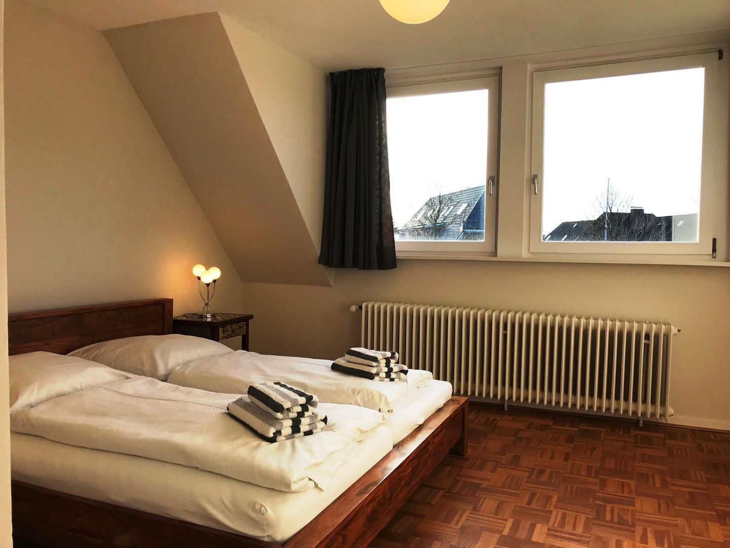 A bedroom with two beds, a radiator, a window with curtains, and a lamp on a bedside table.