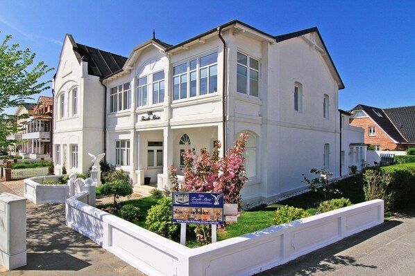 A white two-story building with a balcony, a signboard, and a well-maintained garden.