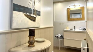 Un baño moderno con un cuadro de temática náutica, un tocador redondo, un espejo con marco de madera y un perchero para toallas.