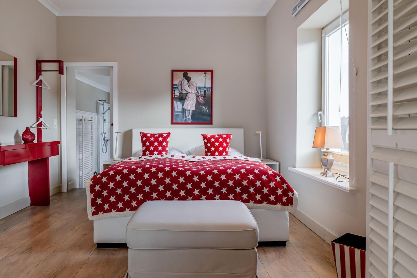 Un dormitorio con cama, mesita de noche, lámpara colgante, ventana con persianas y colcha de cama con estampado de estrellas rojas y blancas.