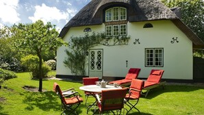 A thatched-roof house with a garden patio, a table with chairs, and a small tree.