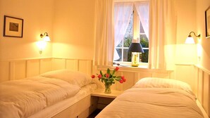 A hotel room with two single beds, a bedside table with a lamp, a window with curtains, and a vase with flowers. - 2. Studio apartment, 65 square meters, 2 bedrooms, sleeps up to 4 people - Weidemannhaus (Sylt-Ost)